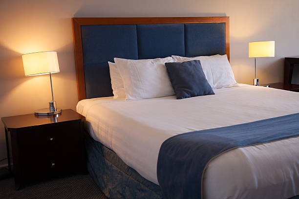 Close up of a bed in a hotel room. The bedside lamps are on. The image features blue colors throughout the decor.